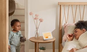 Google Nest Hub på ett bord bredvid en säng där en kvinna ligger och sover, ett litet barn är på väg in genom dörren.