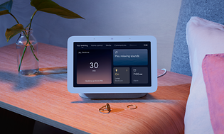 Google Nest Hub på ett nattduksbord, bredvid står en vas med blad och ett par ringar. 