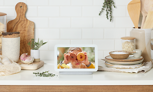 Google Nest Hub på köksbänk bland andra köksredskap. På skärmen syns en flicka med donuts. 