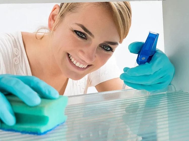 Kvinna städar ur kylskåp med blå diskhandkar på och en tvättsvamp och rengöringsspray i högsta hugg. Bild tagen innifrån kylskåp