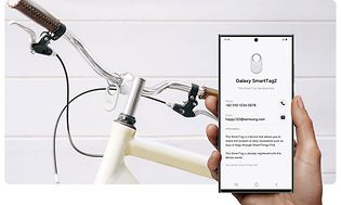 White Samsung SmartTag2 Bluetooth tracker on a bike and hand holding smartphone with App to locate the tracker