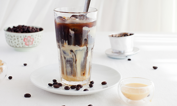 Ett högt glas fyllt med iskaffe på ett vitt bord med kaffebönor utspridda