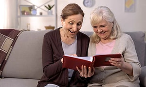 Två kvinnor sitter i en soffa och tittar i ett rött fotoalbum tillsammans. Ena kvinnan är lite äldre. 