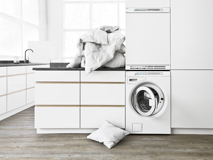 White washing room with front loaded washing machine and a pile of laundry on top of a tabletop above it