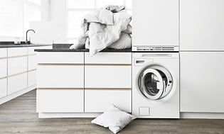 White washing room with front loaded washing machine and a pile of laundry on top of a tabletop above it