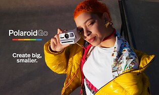 Tjej med orange hår och gul jacka som håller en Polaroid Go-kamera med texten “Create big, smaller.”