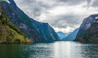 Foto med norsk natur, fjäll och fjordar och några båtar längre bort. Dramatiskt landskap. 