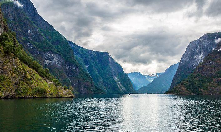 Foto med norsk natur, fjäll och fjordar och några båtar längre bort. Dramatiskt landskap.