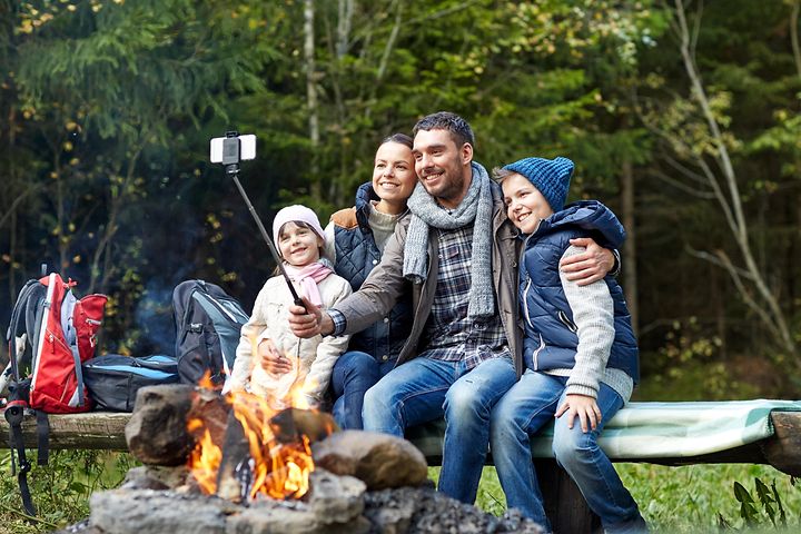 Familj som sitter på en bänk vid en lägereld och tar en selfie, flera ryggsäckar står vid sidan av. 