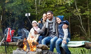 Familj som sitter på en bänk vid en lägereld och tar en selfie, flera ryggsäckar står vid sidan av. 