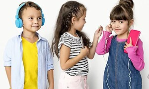 Tre små barn som står vid en vit vägg. En pojke med gul t-shirt och blå hörlurar och två flickor med en iphone och hörlurar. 