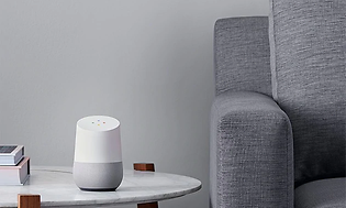 Grå/vit Google home enhet placerad i vardagsrummet, på ett bord bredvid en fåtölj.
