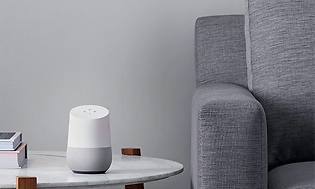 Grå/vit Google home enhet placerad i vardagsrummet, på ett bord bredvid en fåtölj. 