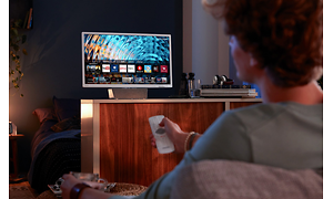 TV på avstånd i ett mörkt rum med sovrum bakom, en man sitter med fjärrkontroll i handen och försöker fixa bildinställningarna.