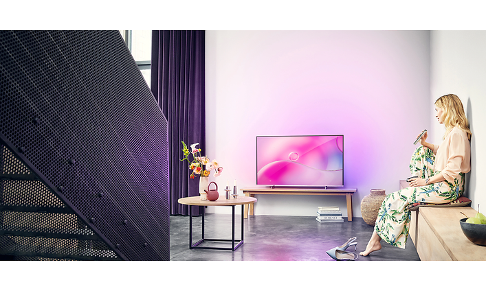 TV som sprider rosa/lila ljus i rummet. Trappa kommer ner till vänster i bild och till höger mot väggen sitter en kvinna.