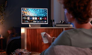 En man sitter i soffan med en vit fjärrkontroll riktad mot TV:n en bit bort, bakom TV:n döljer sig ett sovrum.