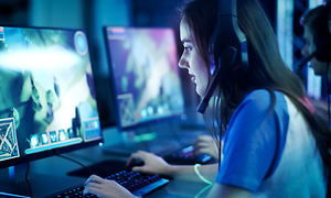 En tjej med headset på sitter och gamar i en spelhall med många andra datorer runt omkring.