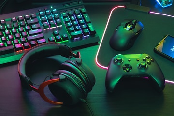 Detaljbild på ett gaming headset bredvid en xbox kontroll och tangentbord med mus. Mörkt rum upplyst av neon färger. 
