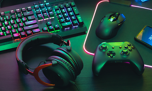 Detaljbild på ett gaming headset bredvid en xbox kontroll och tangentbord med mus. Mörkt rum upplyst av neon färger.