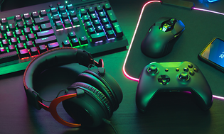 Detaljbild på ett gaming headset bredvid en xbox kontroll och tangentbord med mus. Mörkt rum upplyst av neon färger. 