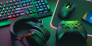 Detaljbild på ett gaming headset bredvid en xbox kontroll och tangentbord med mus. Mörkt rum upplyst av neon färger. 