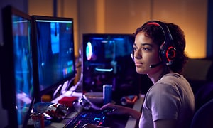 En tjej sitter och spelar i ett gaming rum med många datorer. Hon har på sig ett headset med mikrofon och rött ljus. 