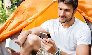 Man sitter utanför ett orange tält och håller i en mobiltelefon och en vit trådlös högtalare. 
