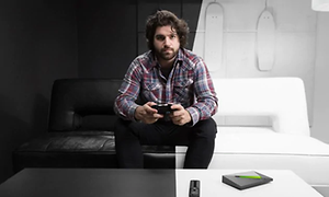 Man med spelkontroll i händerna sitter i en soffa i ett rum som är hälften svart, hälften vitt. Nvidia Shield ligger på bordet.