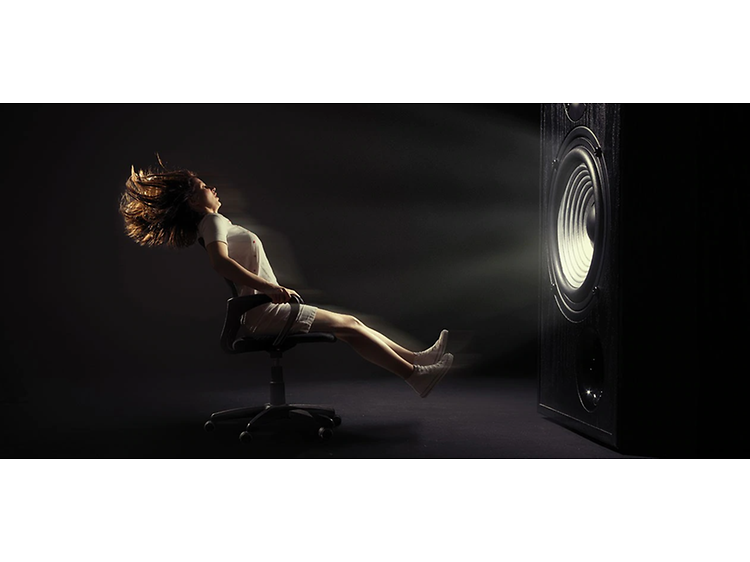 En kvinna som sitter på en kontorsstol med hjul i ett mörkt rum framför gigantisk subwoofer och är på väg att blåsa bort.