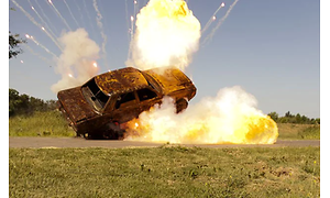 En bil på en grusväg mitt ute på ett fält som exploderar. Bilen har lyft någon meter ovan marken i explosionen.