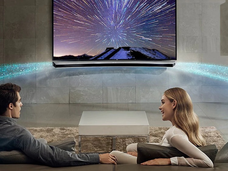 Ett par sitter i TV-soffan i ett modernt vardagsrum med surroundsystem. Ljus symboliserar ljudet som sprider sig ut i rummet. 