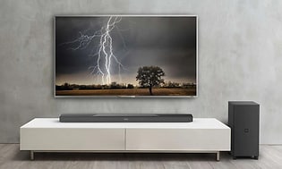 Stor TV uppsatt på väggen med bild på en åskblixt. Tillhörande högtalare vid sidan och soundbar placerad på en vit TV-bänk.