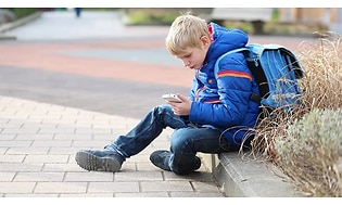 Ett barn med ryggsäck sitter på en trottoar och använder sin mobiltelefon.