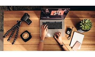 Bild ovanifrån på en person som redigerar video på en Macbook. Vid sidan ligger en kamera med stativ, en mobil och ett block. 