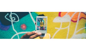 Man som står framför en färgglad vägg och håller upp en polaroidbild, på sig själv, framför ansiktet. 