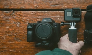 En svart kompaktkamera och actionkamera med tillhörande fäste placerat på ett bord i trä. 