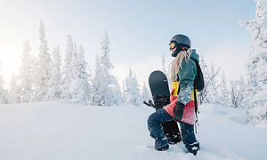 Kvinna som pulserar genom snön i ett vackert vinterlandskap med en snowboard under armen.