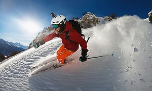 Man som åker skidor på väg nedför ett berg, pudersnö skvätter upp bakom. 