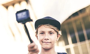 En pojke sträcker upp en actionkamera som sitter fast på en selfie-pinne, och poserar för att ta en bild av sig själv. 