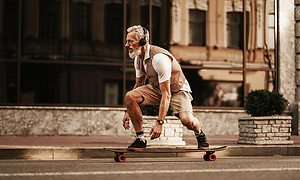 En gammal man med vitt hår och skägg åker skateboard på en gata, med hörlurar på huvudet.