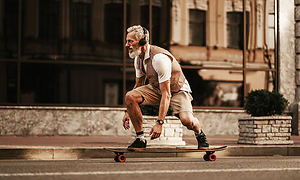 En gammal man med vitt hår och skägg åker skateboard på en gata, med hörlurar på  huvudet.