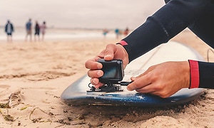 Man som fäster en actionkamera på en surfbräda som ligger på stranden. I bakgrunden syns vattnet och folk som står på stranden.