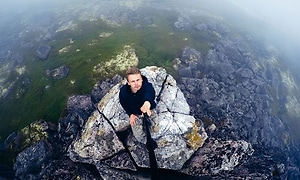 Man som klättrat upp på något slags stenrös sträcker upp en actionkamera på en lång pinne för att ta en cool bild. 