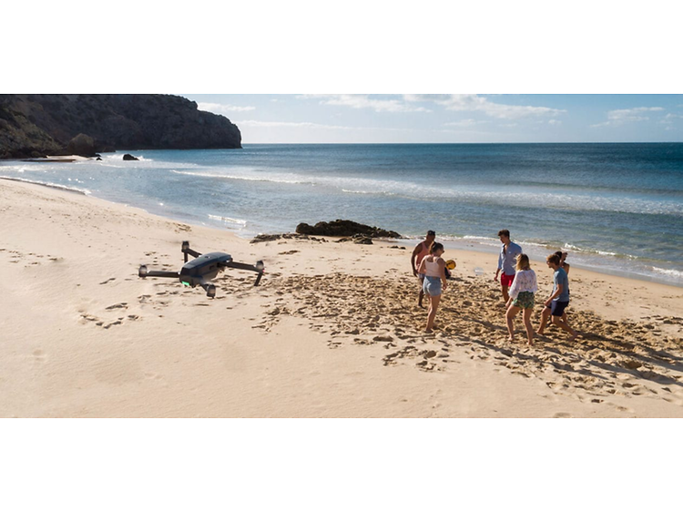 Fem personer spelar boll på en strand, till vänster synns en svart drönare som filmar händelsen ovanifrån.