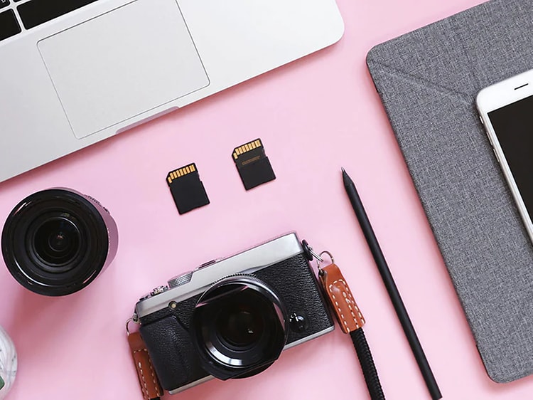 En kamera, ett objektiv, två olika minneskort, en penna, en iPhone och en Macbook på ett rosa bord. 