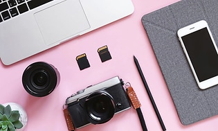 En kamera, ett objektiv, två olika minneskort, en penna, en iPhone och en Macbook på ett rosa bord. 