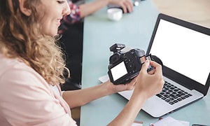 Kvinna sätter i ett minneskort i en systemkamera. En macbook står i bakgrunden på ett bord. 