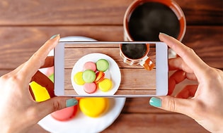 Kvinna som tar ett foto med sin mobiltelefon av färgglada makroner och kaffe på ett träbord.