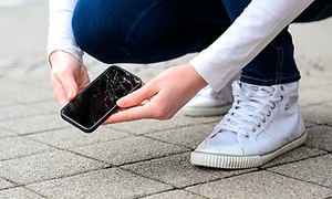 Kvinna i jeans och vita sneakers plockar upp en svart mobil från marken som har krossad skärm. 