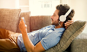 Man ligger på en soffa och ser på mobiltelefonen med vita hörlurar inkopplade.